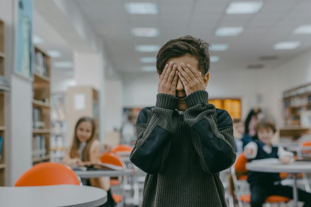Young boy covering his face in a classroom while other students watch and laugh.
