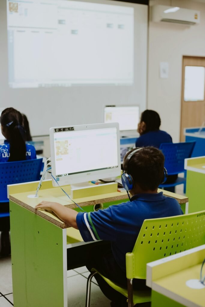 A child wearing headphones focuses on a computer screen in a brightly lit classroom.