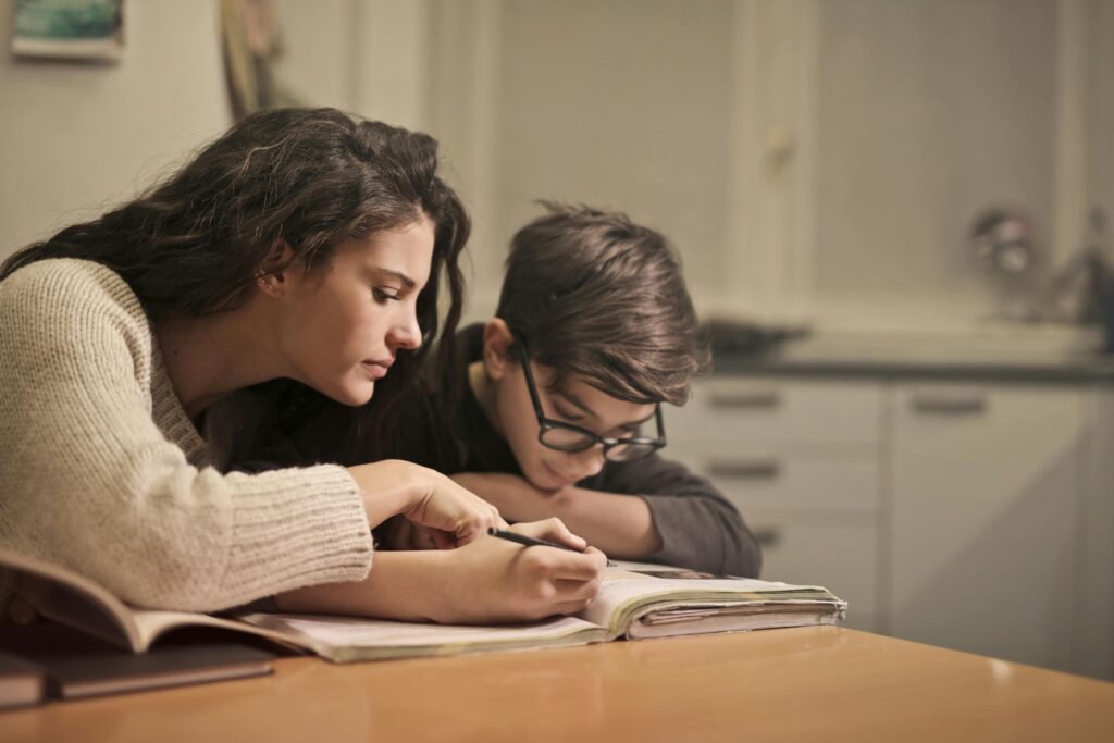 A parent and child sitting at a table, working on homework together with books, notebooks, and a laptop, showcasing a supportive learning moment.